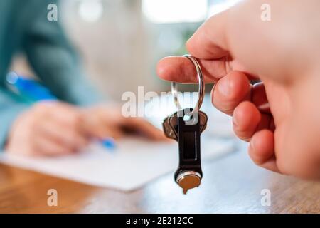 Die Schlüssel hält die Hand eines Mannes. Im Hintergrund, in einer Unschärfe, signiert eine Person Dokumente. Nahaufnahme der Hände. Vermietung und Kauf von Immobilien. Stockfoto