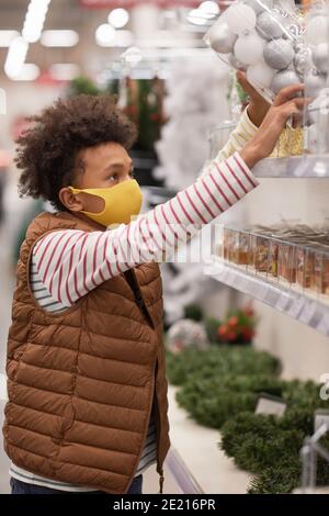 Seitenansicht Porträt von Jugendlichen afroamerikanischen Jungen mit Maske in Supermarkt während Erreichen Weihnachtsdekorationen auf dem obersten Regal Stockfoto