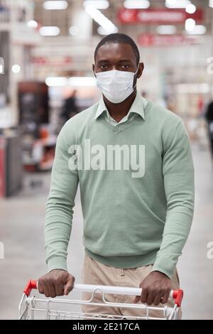 Vertikales Porträt des afroamerikanischen Mannes mit Maske im Supermarkt und Blick auf die Kamera, während Sie den Einkaufswagen schieben Stockfoto