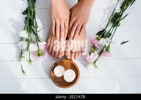 Draufsicht auf weibliche Füße und Hände mit pastellrosa Emaille auf den Nägeln neben den Blumen und den Kerzen auf dem weißen Holz Oberfläche Stockfoto