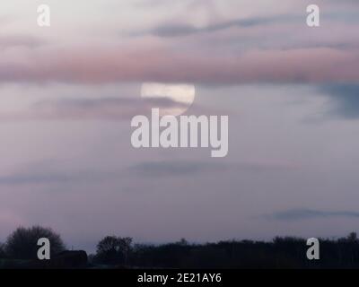 Der Vollmond hinter einer violetten Wolke bei Sonnenuntergang im St Aidans Nature Reserve in Leeds, West Yorkshire an einem kalten Wintertag. Stockfoto