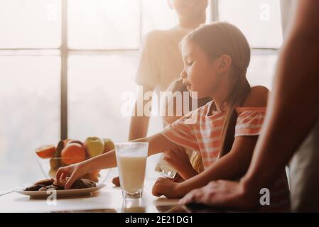 Cropped Bild von Gay paar mit ihren angenommen niedlichen Töchter Kochen auf Küche. LGBT-Familie zu Hause. Kleines Mädchen essen Kekse und Milch trinken. Stockfoto