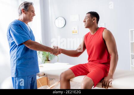 Glücklicher afroamerikanischer Mann in Sportswear, der mit Chiropraktiker die Hände schüttelt In der Klinik Stockfoto