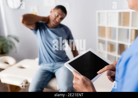 Reifer Arzt mit digitalen Tablette in der Nähe afroamerikanischen Patienten leiden Von Nackenschmerzen auf verschwommenem Hintergrund Stockfoto