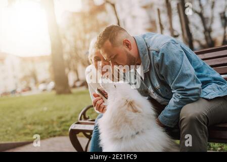 Junge glücklich lächelnde Frau und Mann streicheln ihr Weiß Flauschiger Hund, während er auf einer Gassenbank sitzt Stockfoto