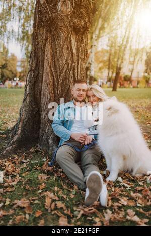 Glückliche hübsche Frau und schöner Mann posiert mit weißen Pelz Spielerischer Hund samoyed Rasse beim Sitzen in der Nähe von großen Baum Herbstpark Stockfoto