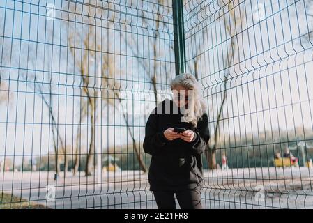 Taille von jungen hübschen Dame in der Nähe des Basketballs stehen platz und tragen stilvolle Kleidung während der Verwendung des Telefons Stockfoto