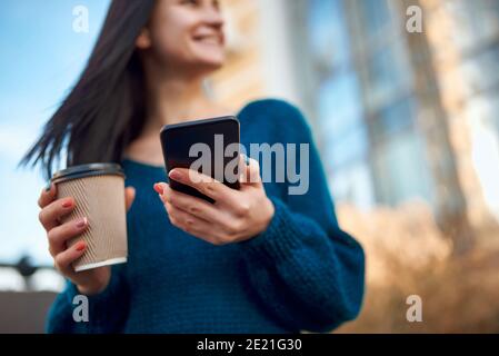 Nahaufnahme eines schwarzen Smartphones und Einwegglases Von Kaffee in den Händen einer verschwommenen lächelnden jungen Frau Stockfoto