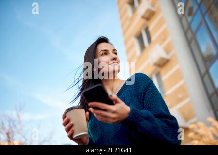 Junge schöne Frau, die durch hohe moderne Gebäude schlendert und hält Handy und Tasse Kaffee in den Händen Stockfoto