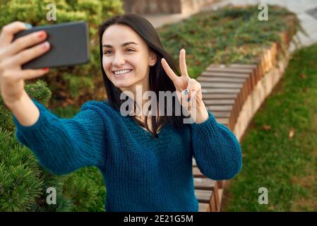 Trendige junge Frau, die ein positives Selfie auf einem Gehweg eines grünen Blumenbeds macht, glänzendes Lächeln und Siegeshand zeigt Stockfoto