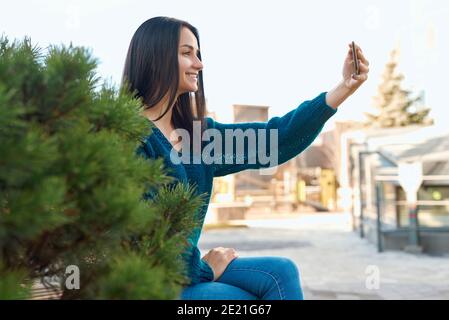 Seitenansicht einer lächelnden jungen Frau, die ein Selfie aufschießt Ihr Handy in der Nähe einer schönen dicken Zwergkiefer Stockfoto