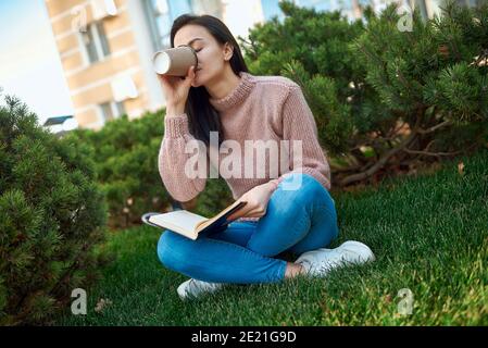 Charmante junge Dame trinken Latte aus einem Papierglas und Setzen Sie das Copybook, während Sie auf grünem Gras innen sitzen Ein Türgarten Stockfoto