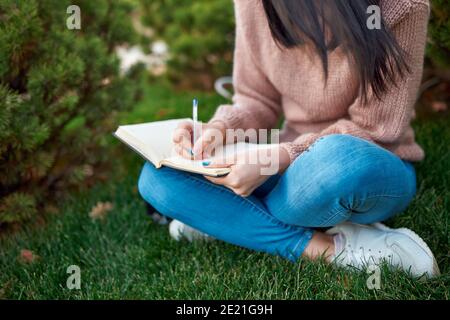 Nahaufnahme einer jungen Frau in Jeans und Pullover Notizen in ihrem Papierplaner machen, während sie darauf sitzen Gras im Freien Stockfoto
