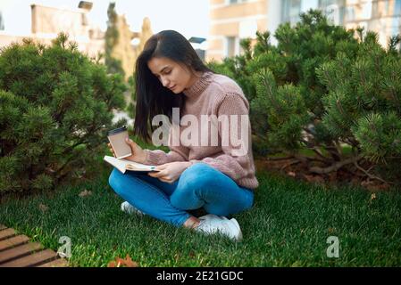 Attraktive konzentrierte junge Frau mit interessanten Buch sitzend mit gekreuzten Beinen auf Ein Gras außerhalb der modernen Stadtbauten Stockfoto