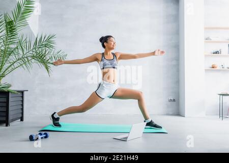 Fröhliche afroamerikanische Frau in Sportkleidung üben Krieger Pose in der Nähe Laptop zu Hause Stockfoto