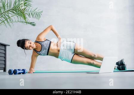 Happy african american Sportlerin Ausübung in Seitenplanke Pose auf Fitnessmatte in der Nähe von Laptop und Hanteln Stockfoto