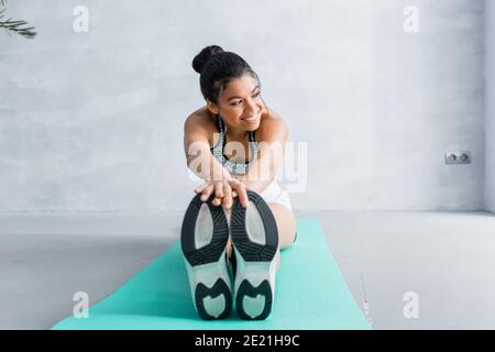 Glückliche afroamerikanische Frau, die weg schaut, während sie vorwärts sitzt Bend Übung auf Fitness-Matte Stockfoto