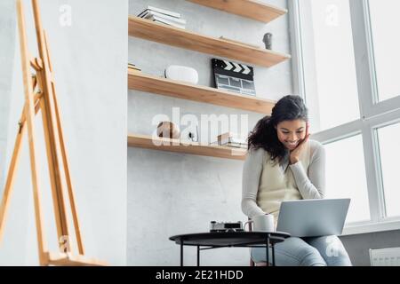 Lächelnde afroamerikanische Frau mit Laptop zu Hause in der Nähe Staffelei Bei unscharfem Vordergrund Stockfoto