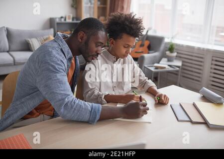 Porträt von fürsorglichen afroamerikanischen Vater hilft Sohn Hausaufgaben oder Studieren während zusammen am Schreibtisch sitzen in minimalem häuslichen Interieur Stockfoto