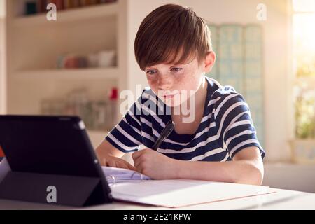 Junge Sitzt Am Küchentisch Tun Hausarbeit Mit Digital Tablet Stockfoto