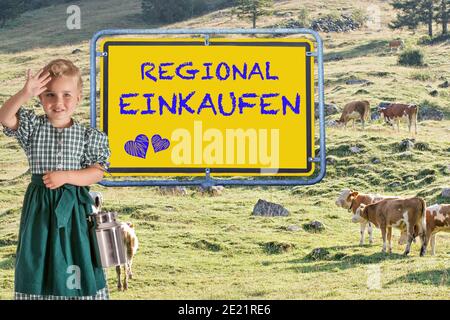 Ein kleines Kind in einem Dirndl mit einem Milchkännchen In der Hand vor einer Almwiese mit Kühe und das Schild 'Buy regional'.Beschriftung Deutsch Stockfoto