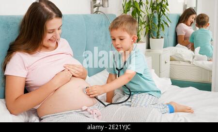 Kleiner Junge hört Mütter schwanger großen Bauch mit Stethoskop. Konzept der Gesundheitsversorgung und medizinische Untersuchung während der Schwangerschaft Stockfoto