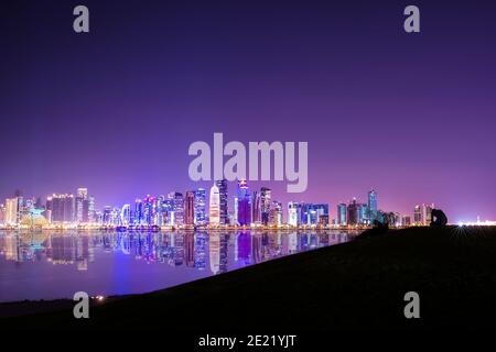 (Selektiver Fokus) atemberaubender Panoramablick auf die Skyline von Doha, beleuchtet in der Abenddämmerung in der Ferne und einige Leute entspannen sich auf einem Hügel im Vordergrund Stockfoto