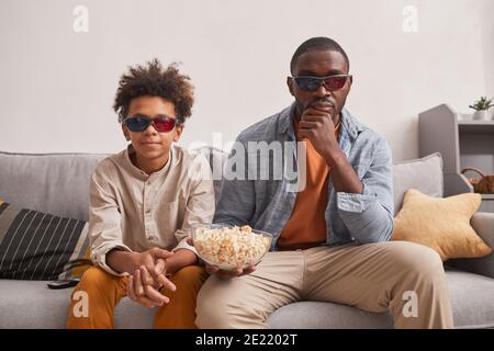 Moderner afroamerikanischer Familienmensch und sein jugendlicher Sohn sitzen Auf dem Sofa im Wohnzimmer 3D-Filme ansehen und essen Popcorn Stockfoto