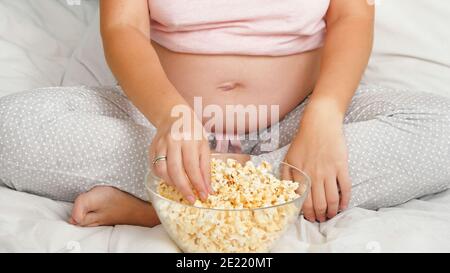 Nahaufnahme der hungrigen schwangeren Frau im Schlafanzug auf dem Bett sitzen Und Popcorn essen Stockfoto