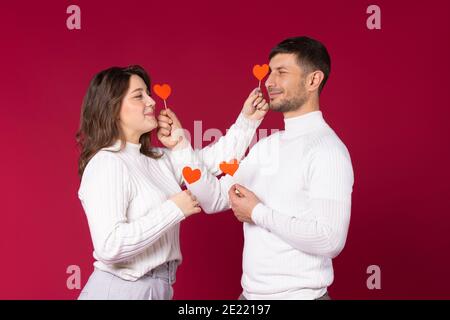 Ein liebevolles Paar posiert mit roten Papierherzen auf rotem Hintergrund. Verbringen Sie Zeit am Valentinstag. Stockfoto