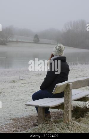 Eine Frau, die sich auf einer frostbedeckten Holzbank mit Blick auf einen See im Winter zurück zur Kamera sitzt. Speicherplatz kopieren. Stockfoto