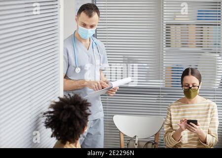 Porträt des männlichen Arzt trägt Maske und hält Zwischenablage während Im Gespräch mit den Patienten, die in der medizinischen Klinik Schlange stehen Stockfoto