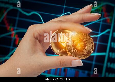 Makroökonomische weibliche Hand, die Bitcoin und ethereum nebeneinander vor dem Börsenchart hält. Stockfoto