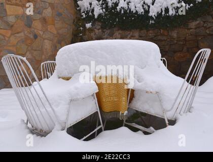Tisch mit orange-gelber Tischdecke und zwei Stühlen mit Schnee. Gartenszene nach dem Durchzug des Sturms Filomena durch Spanien. Januar 2021. Stockfoto