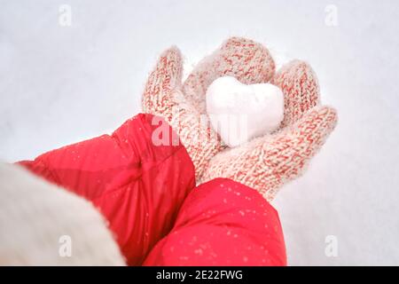 Frauen Hände in roten Fäustlingen halten ein Herz aus Schnee, Nahaufnahme. Frau hält einen herzförmigen Schneeball für valentinstag Stockfoto