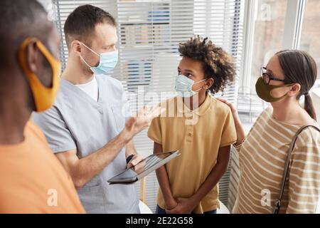 Porträt eines männlichen Arztes, der mit der Familie spricht, während er im Wartezimmer im Krankenhaus steht und alle Masken trägt Stockfoto