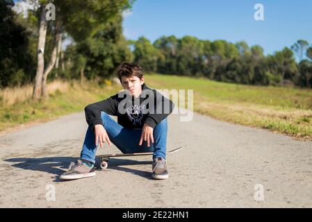 Hübscher Teenager-Mann mit einem Skateboard auf Asphaltstraße sitzen Stockfoto