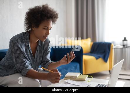 Schwarze Frau, die elektronische Rechnungen auf dem Mobiltelefon analysiert, während sie verwaltet Finanzen Stockfoto