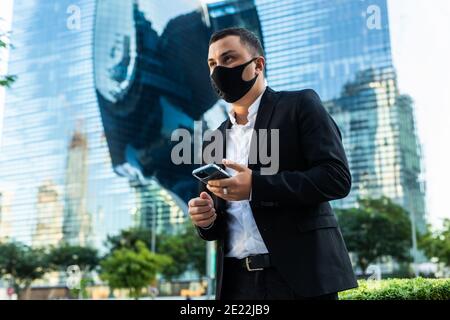 Kaukasischer junger schöner Mann in medizinischer Maske Klopfen und Scrollen auf Handy außerhalb im Business Center. Glücklicher Geschäftsmann im Atemschutz Stockfoto