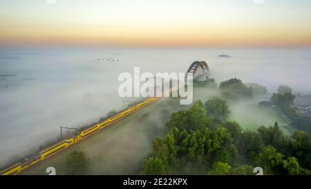 Zweidecker-Intercity-Zug von Nederlandse Spoorwegen durch dichten Nebel in Richtung Eisenbahnbogenbrücke, Gelderland, Niederlande Stockfoto