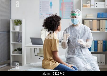 Porträt eines Arztes mit voller Schutzausrüstung, der einen Covid-Abstrichtest macht, während er einen afroamerikanischen Jungen in der Klinik untersucht, Raum kopieren Stockfoto