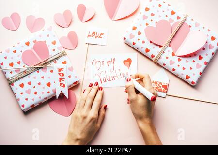 Elegante weibliche Hände mit roter Maniküre schreiben eine Notiz SEI MEIN VALENTINSTAG mit einem Filzstift. Eingewickelte Geschenke auf dem Tisch und viele Herzen. Stockfoto