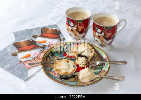 Teller mit Mince Pies, eine mit Puppe Sahne aufgeschnitten, auf dem Tisch neben zwei Tassen cremigen Kaffee und Servietten. Süße Leckerbissen zur Weihnachtszeit, lecker Stockfoto