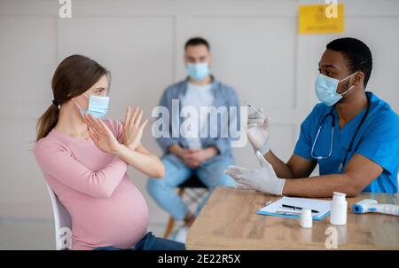 Schwangere Frau in Gesichtsmaske erschrocken, um Coronavirus-Impfstoff-Injektion zu machen, machen STOP-Geste in der Klinik, Panorama Stockfoto