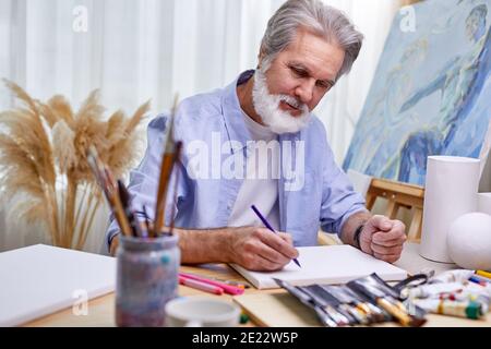 Maler zeichnet zu Hause im hellen Raum, graubärtiger Mann schafft ein Meisterwerk mit einem Bleistift Stockfoto
