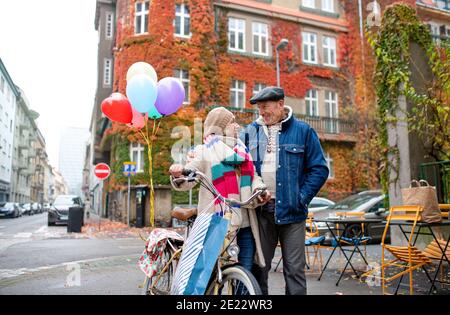 Glückliches Seniorenpaar mit Fahrrad und Luftballons draußen auf der Straße in der Stadt. Stockfoto