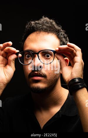 Mann mit Brille starrt in einem schwarzen Poloshirt in die Kamera, während er seine Brille vom Gesicht hält. Schwarzer Hintergrund. Optisches Konzept Stockfoto