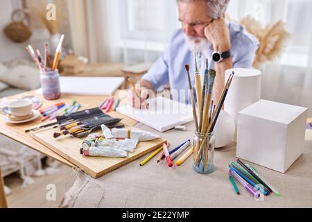 Pinsel in Glasdosen und Gouache, Farben, Bleistifte auf dem Tisch und nachdenklicher Maler im Hintergrund. Kunstkonzept Stockfoto