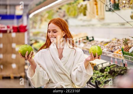 Frau wählt frisches Gemüse und Obst im Laden, trägt Bademantel. Junge Frau Lebensmittel im Supermarkt kaufen Stockfoto