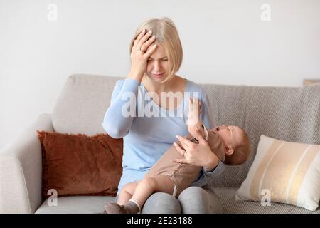 Erschöpft blonde Frau mit Migräne, hält kleines Baby, Zuhause innen Stockfoto